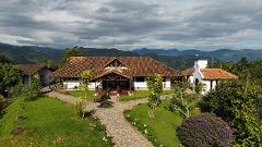 A charming rural estate with a large house, a smaller building with a bell tower, and a lush garden set against a backdrop of rolling green hills and a cloudy sky.