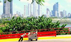 Panama City and Darien Gap A man in a hat pushes a food cart up a yellow path with a red railing, with the Panama City skyline in the background.