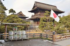 A scenic view of a traditional Japanese building complex with a prominent Japanese flag, surrounded by lush greenery, and a display of sake barrels.