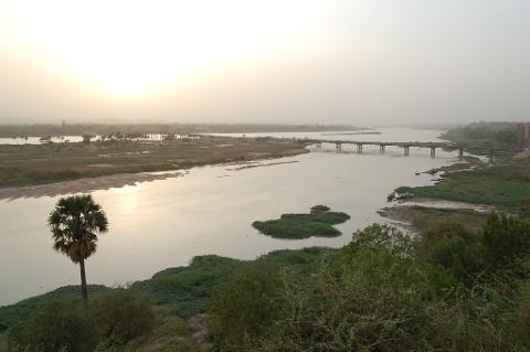 An aerial shot of a wide, muddy river with lush green vegetation and a bridge stretching across, all bathed in the soft light of a hazy sunset.