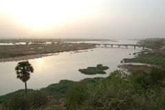 An aerial shot of a wide, muddy river with lush green vegetation and a bridge stretching across, all bathed in the soft light of a hazy sunset.