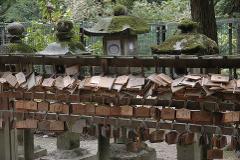 A wooden fence adorned with numerous wooden plaques, some stacked haphazardly, in front of moss-covered stone lanterns.