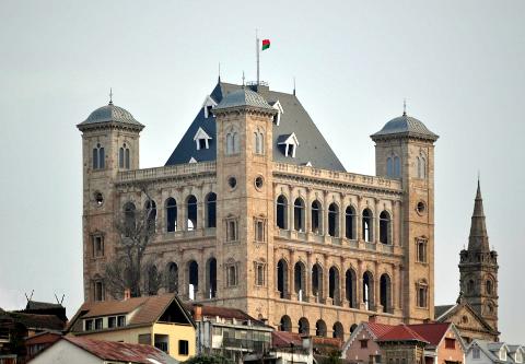 Overlooking a cityscape, the Rova of Antananarivo features a stone structure with arched windows and a flag atop a gray roof, juxtaposed with a neighboring church tower.