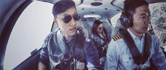 Three people are in the cockpit of a small plane, possibly a helicopter. One of them is wearing a pilot's uniform and headphones, suggesting he may be the pilot.