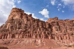 A towering red rock formation rises against a blue sky with scattered white clouds, showcasing a rugged, layered landscape.