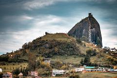 A striking landscape featuring the massive El Peñol rock formation with a stairway, set amidst lush green hills and scattered buildings under a cloudy sky.