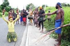 Certainly! Here's a description of the image:A group of people in traditional attire, some with painted bodies and carrying spears/bows, are standing on a road in Papua New Guinea, possibly during the Rabaul Mask Festival.