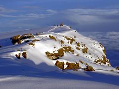 A snow-covered mountain landscape with rocky outcrops, a visible path leading to a group of people at the summit, under a bright blue sky with scattered clouds.