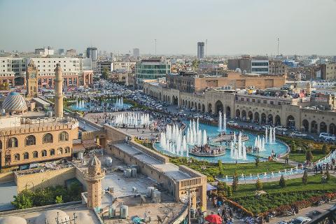 An aerial view showcases a bustling city with fountains and greenery amid historical buildings and modern structures under a clear sky.