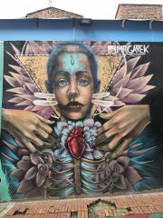 This is a street art mural featuring a person with a heart inside the ribs. The person's face has a blue mark on their forehead, surrounded by geometric and floral elements.
