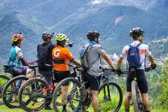 A group of five cyclists pause on a grassy hillside, their mountain bikes beside them, as they admire the scenic view of rolling green mountains.