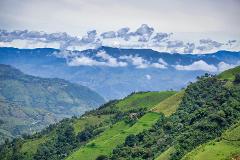 Lush green mountains rise against a backdrop of distant, blue-hued peaks and a sky dotted with white, puffy clouds.