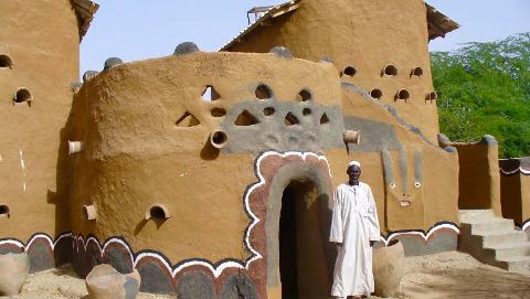 A man stands before a mud-brick building decorated with various geometric shapes and human-like features, under a clear blue sky.