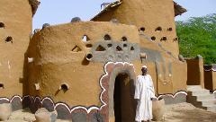 A man stands before a mud-brick building decorated with various geometric shapes and human-like features, under a clear blue sky.