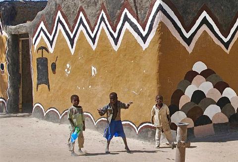 Three young children stand and play in front of a vibrantly painted mud house decorated with geometric patterns and symbolic imagery.