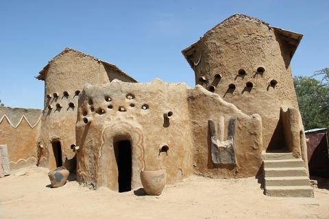 This image shows a traditional mud-brick building with rounded towers and circular openings, featuring a decorative mask on the facade.