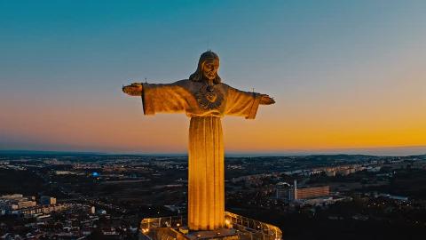 An aerial shot of the Cristo Rei statue illuminated against a sunset sky, overlooking a sprawling urban landscape.