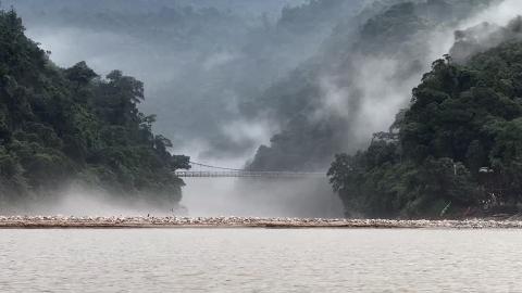 A misty landscape with a bridge connecting tree-covered hills, seen from across a wide river with a sandy, rocky shore.