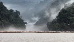 A misty landscape with a bridge connecting tree-covered hills, seen from across a wide river with a sandy, rocky shore.