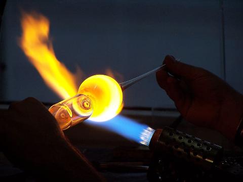 A glassblower shapes molten glass with a torch and tools in a crafting workshop, creating art with fire.