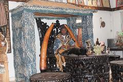 Peuples et civilisations des Grassfields Un homme en tenue traditionnelle est assis royalement sur un trône sculpté orné de défenses, dans une pièce remplie d'objets africains.