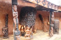 Peuples et civilisations des Grassfields Une collection de statues et d'œuvres d'art en bois africain finement sculptées est exposée contre un mur de couleur terre sous un toit à motifs.