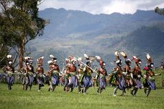 A group of people in traditional costumes and face paint march across a grassy field during the Mount Hagen Festival in Papua New Guinea, with mountains in the background.