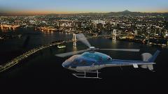 A helicopter flies over a dark body of water with illuminated bridges and a sprawling city skyline in the background.