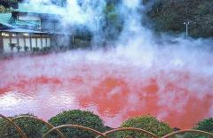 淡い緑色の屋根を持つ日本風の建物の前に、赤い湯気が立つ湖が映っています。