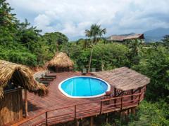 Birding in Colombia - Guajira, Tayrona, and Minca Here's a one-sentence description of the image, keeping it under 320 characters:A tropical resort features a round blue pool on a wooden deck with thatched-roof structures, surrounded by lush greenery and distant mountains under a cloudy sky.