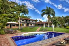 A vibrant image showcases a luxurious outdoor space with a blue-tiled pool in the foreground, a lush green lawn dotted with tables, a traditional-style building with balconies, palm trees, and a clear, blue sky with fluffy white clouds in the background.