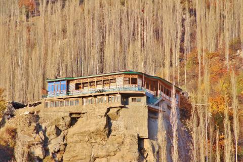 A multi-story building with a green roof sits atop a rock face against a backdrop of tall, thin trees in autumn colors.