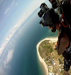 A skydiver is captured mid-air, camera mounted on his helmet, overlooking a stunning coastline where a sandy beach meets the expansive ocean.