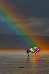 A person stands on a wet beach holding a colorful kite, with a vibrant rainbow arcing across the sky in the background.
