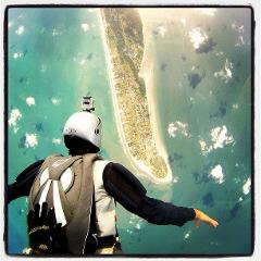 A skydiver with a GoPro helmet camera leaps from a plane, offering a stunning aerial perspective of a coastal island.