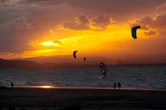 A vibrant sunset over a beach with several people kitesurfing.