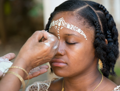 A woman is receiving a white facial decoration on her forehead with intricate patterns, likely as part of a cultural or ceremonial tradition.