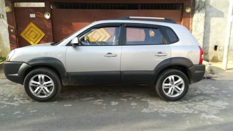 A silver Hyundai Tucson SUV is parked on a road in front of a house with a brown gate.