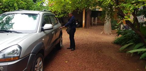 A man in a blue shirt and black pants stands by a silver car, opening the driver's side door. The car is parked on a red gravel driveway surrounded by lush green trees and foliage.