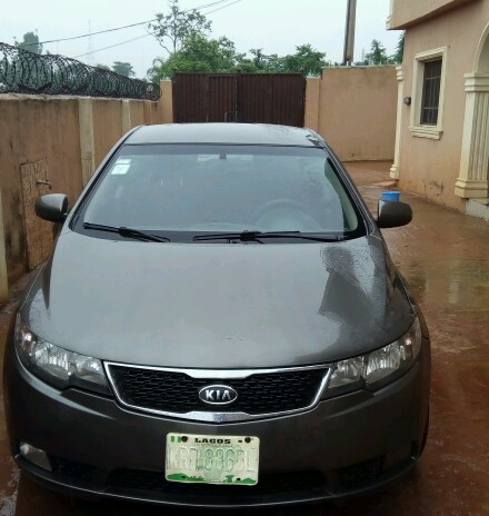 A gray Kia Forte is parked in front of a building with a gated entrance and razor wire along the top of a wall. The license plate reads "LAGOS KRD 886GL".