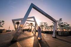 Here's a concise description of the image:A man in traditional attire walks through a series of modern, angular, metallic structures, set against a backdrop of palm trees and a sunset sky, creating a striking contrast of traditional and contemporary elements.