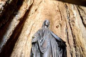 Here's a one-sentence description of the image:A bronze statue of a woman, likely the Virgin Mary, stands with outstretched arms in front of a rough, cavernous rock face.