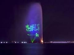 Here's a concise description of the image:A nighttime view of King Fahd's Fountain in Jeddah, Saudi Arabia, illuminated with the Saudi Arabian flag and Arabic script in green light.
