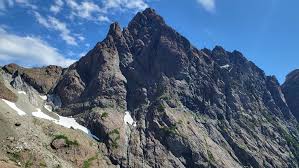 A majestic, rugged mountain with a sharp peak dominates the landscape under a bright blue sky with scattered clouds.