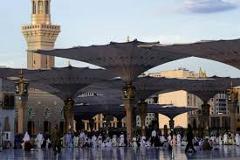 Here's a description of the image:A wide view shows many people praying and walking under large, umbrella-like sunshades surrounding a mosque with a tall minaret in Medina, Saudi Arabia.