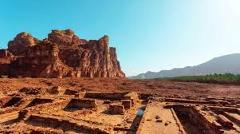 Here's a description of the image:Ancient ruins are foregrounded with a stunning backdrop of red rock formations and a clear blue sky in an arid landscape.