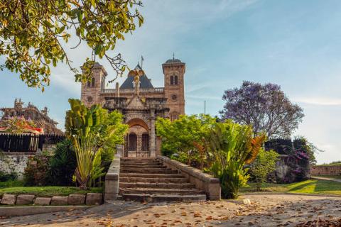 A grand stone building with towers sits behind a staircase and lush green plants under a partly cloudy sky.