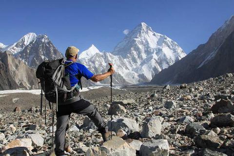 A hiker with a backpack and trekking poles stands on a rocky terrain, gazing at the majestic snow-capped mountains under a clear blue sky.