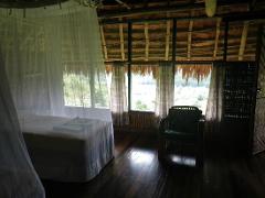 This is an interior shot of Karawari Lodge, showing a room with a bed draped in mosquito netting, a chair, and windows overlooking a green landscape.