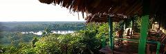 Karawari Lodge (Sepik) A panoramic view from the Karawari Lodge shows a lush rainforest landscape with a river winding through it, seen from a sheltered, elevated porch with thatched roof and rustic seating.